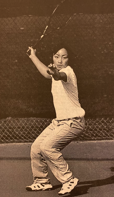 An Asian student in a white t-shirt and light pants is playing tennis on a court. Their stance is focused, holding the racket with both hands, ready to strike. 