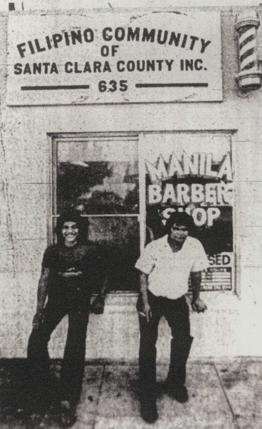 A black and white photo of two men posing in front of a building that says "Manila Barbershop"